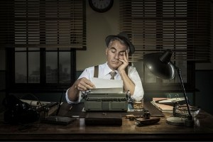 Tired writer at desk at night. All the lights are out so he is just lit by a desk lamp and looks tired and frustrated.