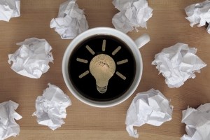 Coffee cup with lightbulb icon in foam surrounded by balled up pieces of paper.